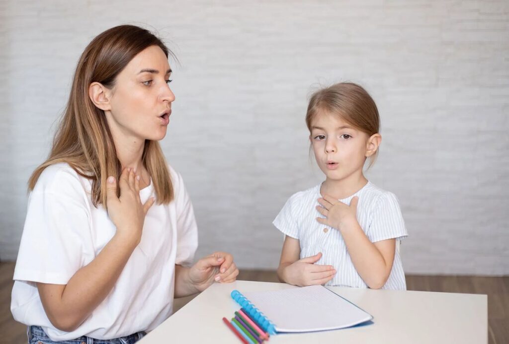 A woman and a young girl sit at a table, smiling as they write in a notebook, enjoying a friendly moment together.
